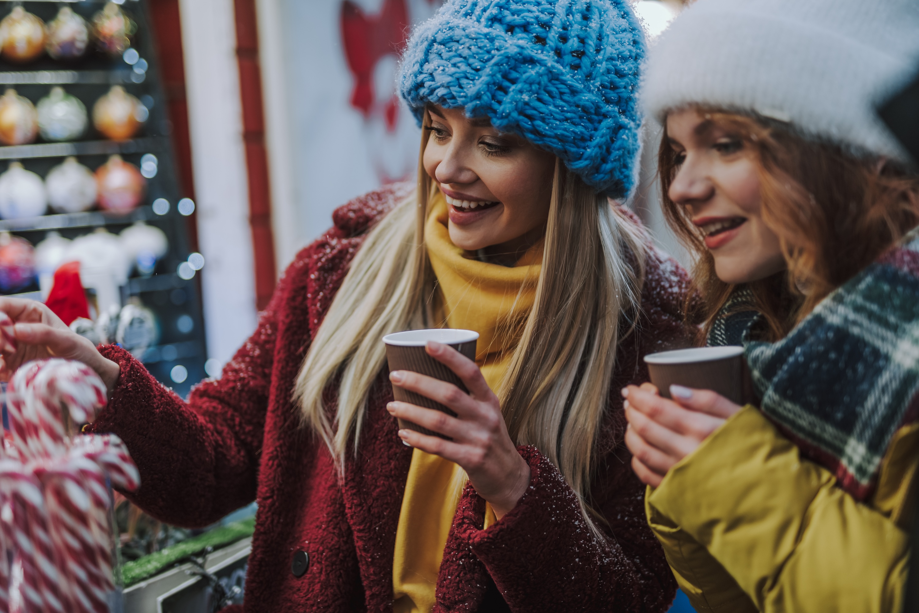 Two smiling women in winter hats and coats holding warm drinks while browsing a festive holiday market with candy canes and ornaments, representing the seasonal community events near The Julian in Sloans Lake, CO.