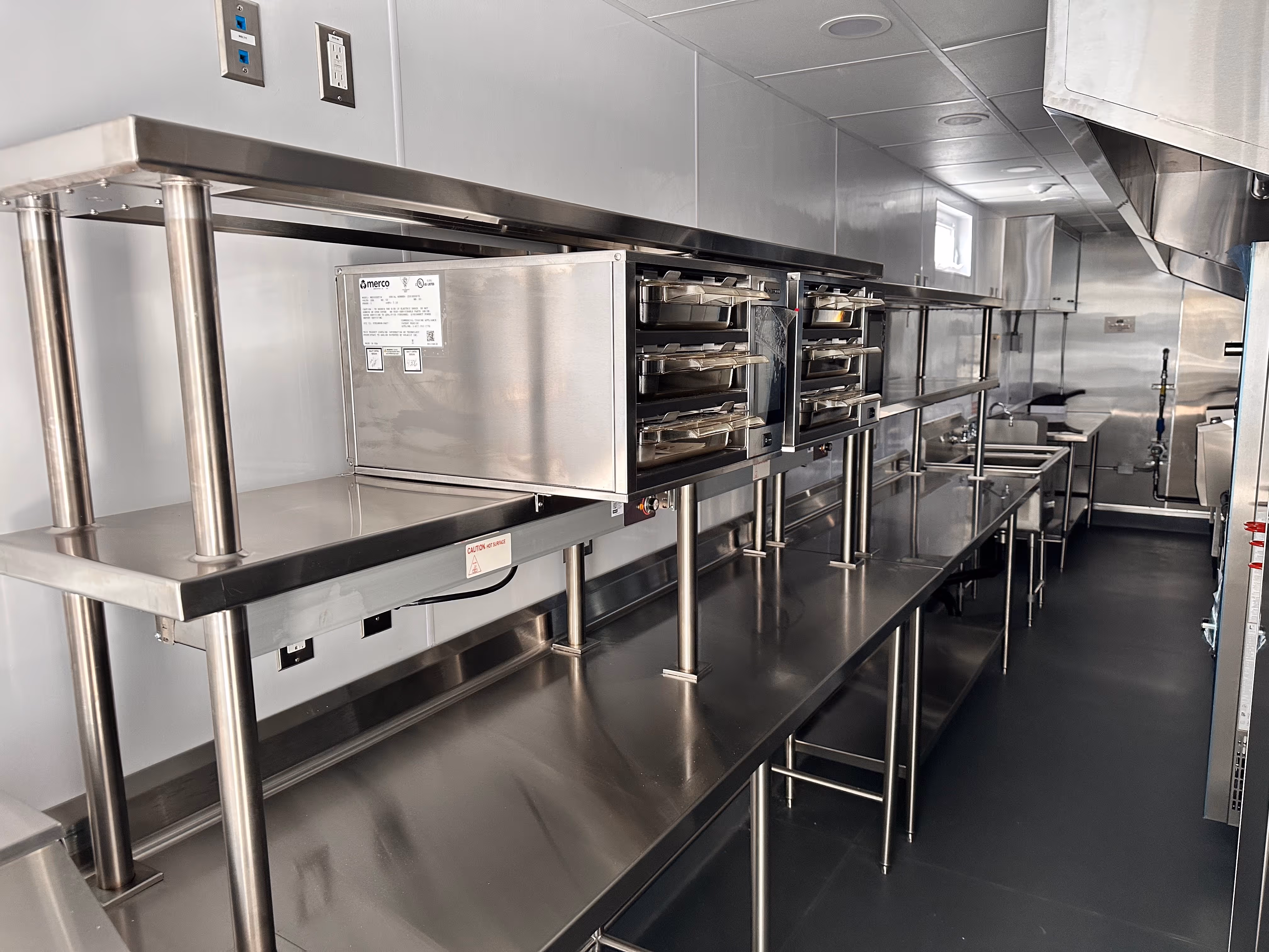 Long narrow commercial stainless steel kitchen with multiple worktables, sinks, and heating trays.