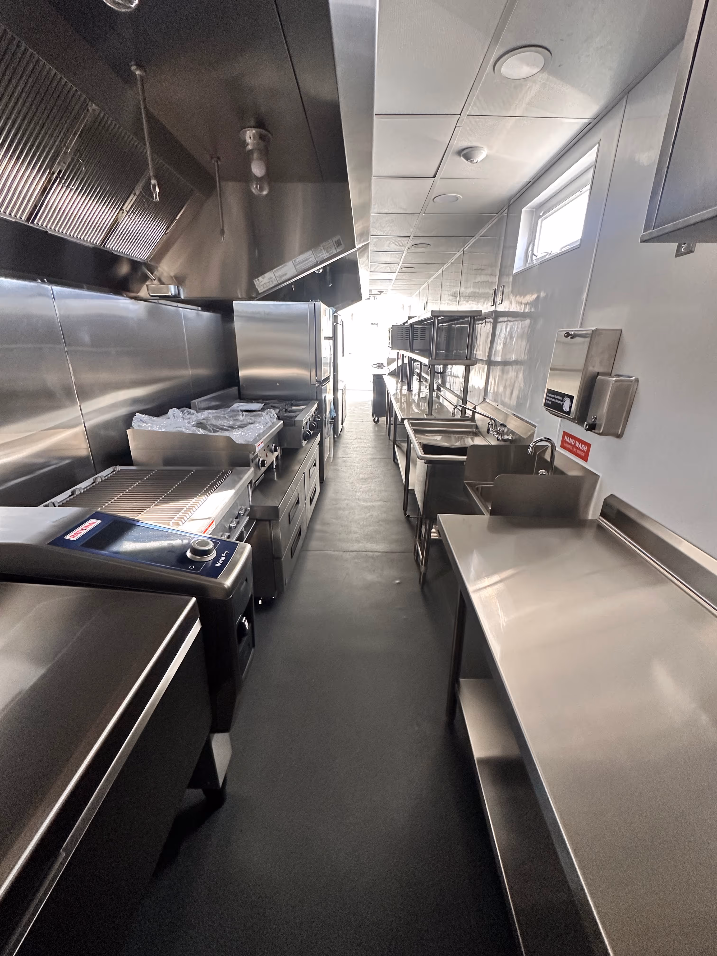 Narrow commercial kitchen with stainless steel appliances, sinks, and preparation tables along both sides.