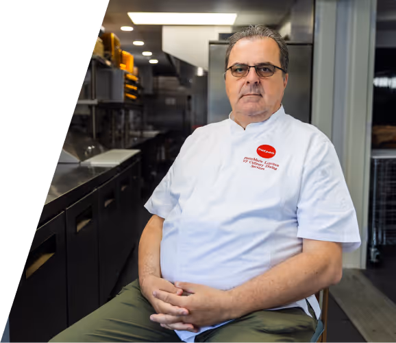Serious male chef wearing glasses and a white uniform sitting in a professional kitchen.