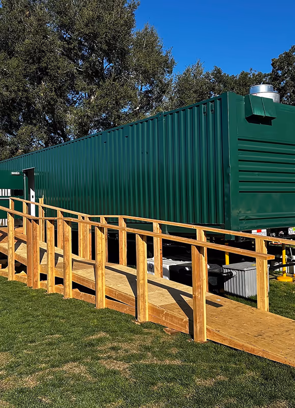 Green metal container building with a wooden wheelchair ramp on a grassy area under a clear blue sky.