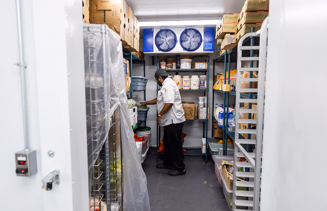 Chef in white uniform arranging fresh produce in a commercial walk-in cooler with shelves stocked with food containers and boxes.