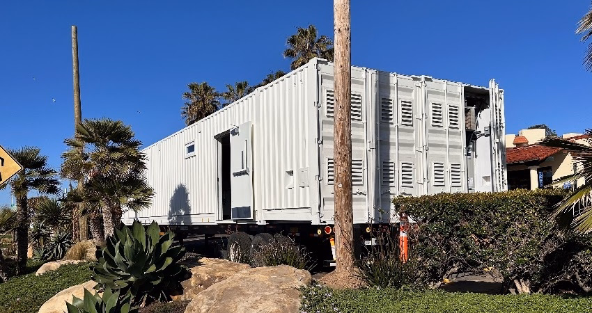 Large white shipping container trailer parked on grass and rocks near palm trees under a clear blue sky.