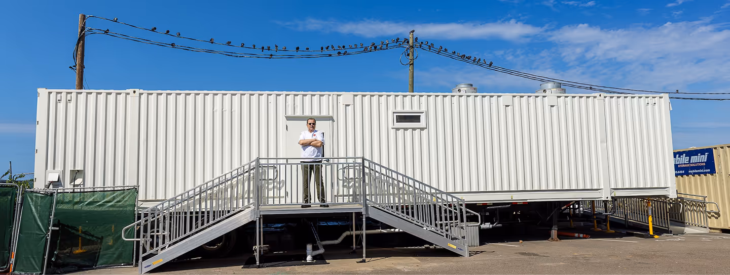 A man stands with arms crossed on a metal staircase platform outside a large white portable office trailer under a blue sky.