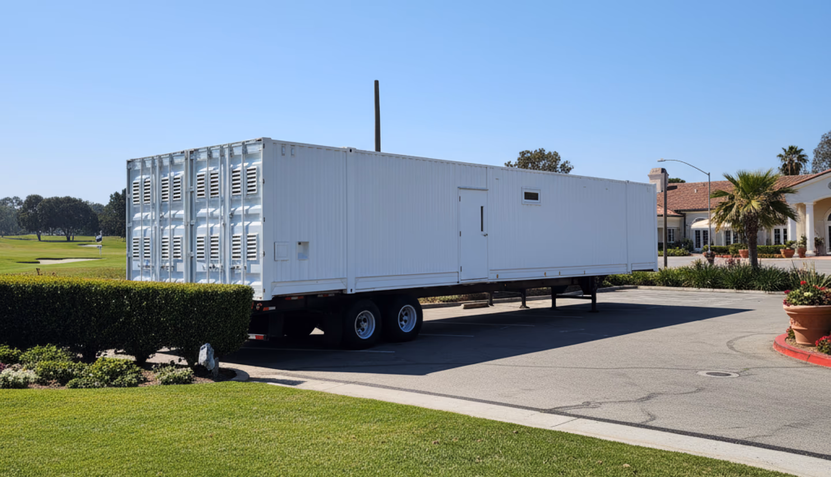 Large white trailer parked on pavement near neatly trimmed bushes and a golf course with a clubhouse in the background under clear blue sky.