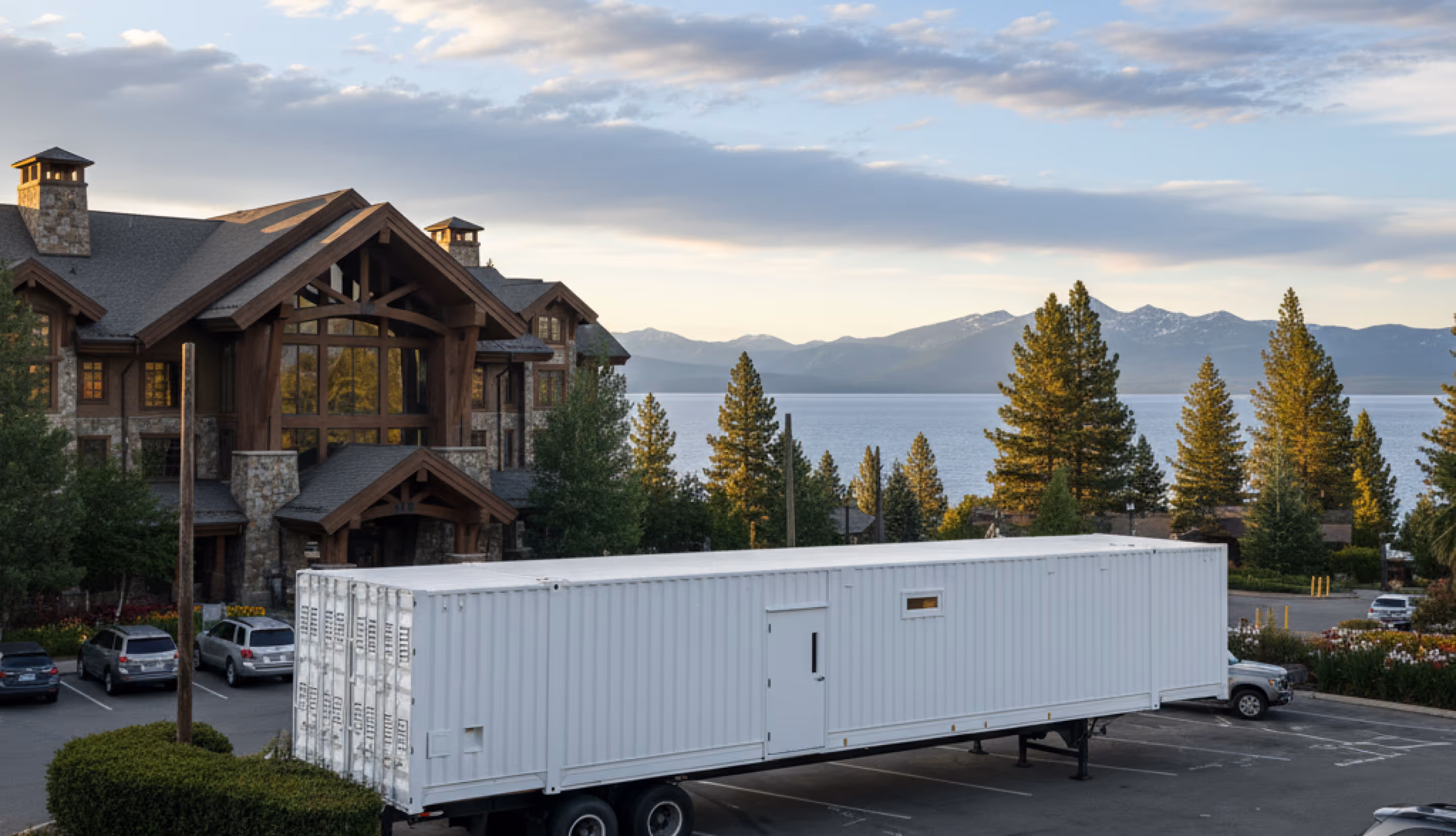Long white trailer parked in a lot near a large stone and wood lodge with pine trees and mountains in the background.
