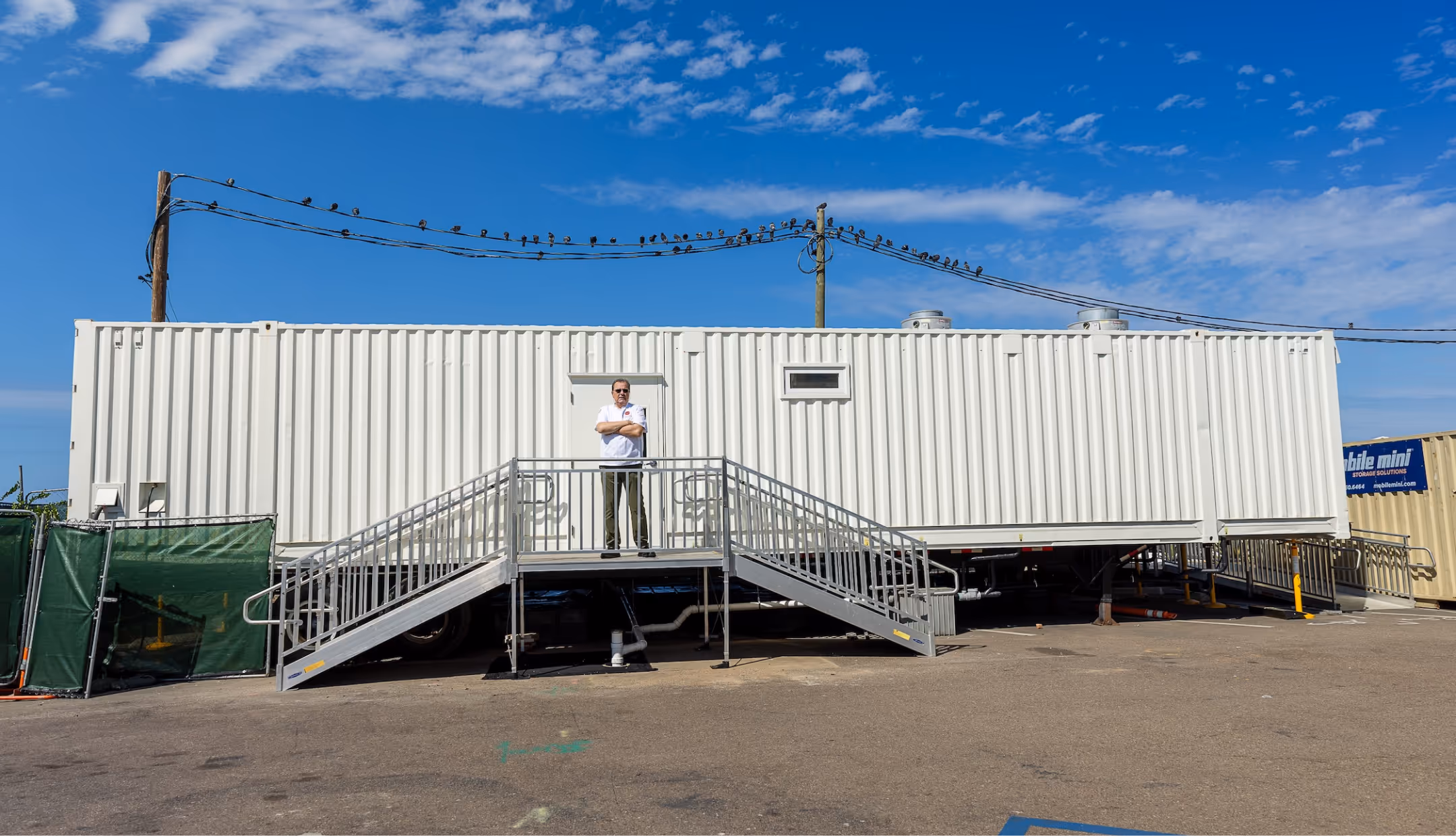 Man standing with arms crossed on metal stairs leading to a white mobile office trailer under a partly cloudy blue sky.