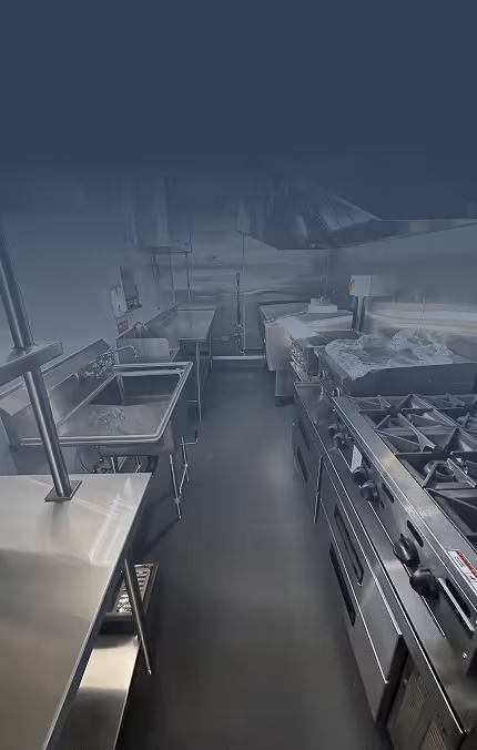 Interior of a commercial kitchen featuring stainless steel countertops, sinks, gas stovetops, and ventilation hood.