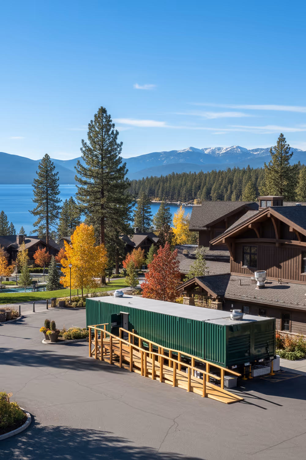 Green mobile kitchen trailer with wooden ramp parked near brown cabins surrounded by trees and mountains with a lake in the background.