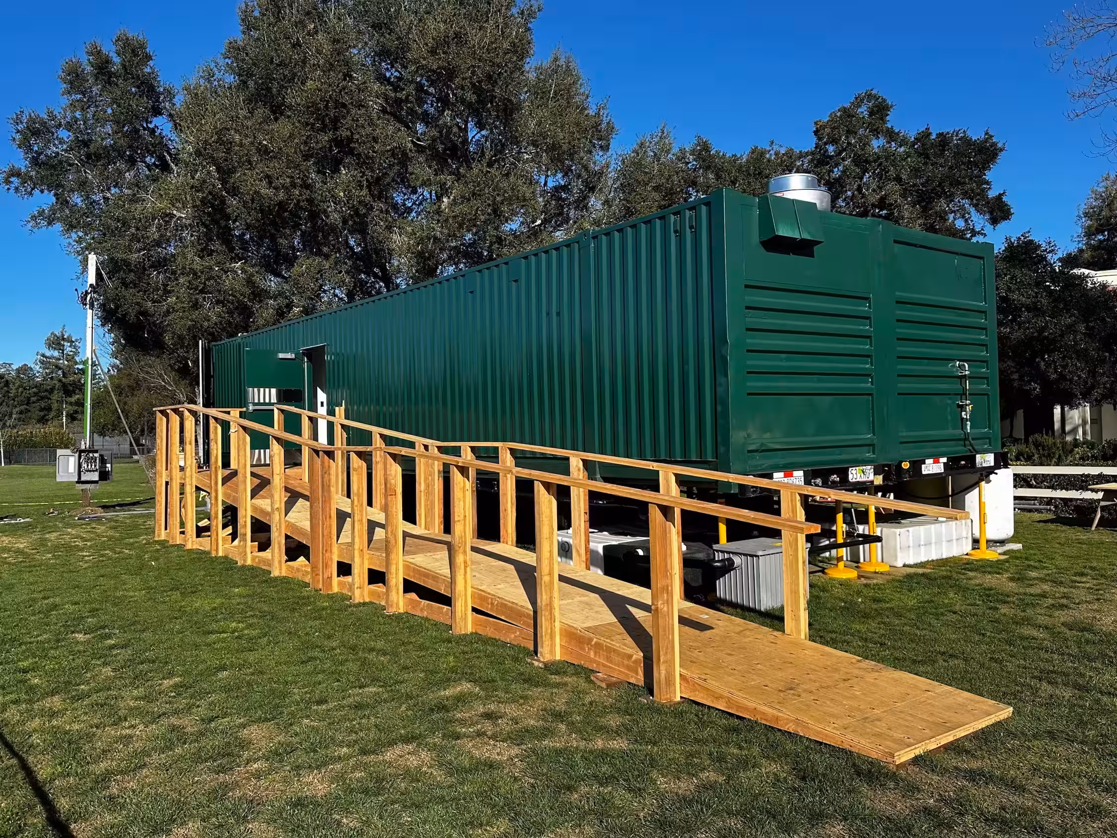 Green mobile kitchen trailer with wooden wheelchair-accessible ramp on a grassy area under a clear blue sky.