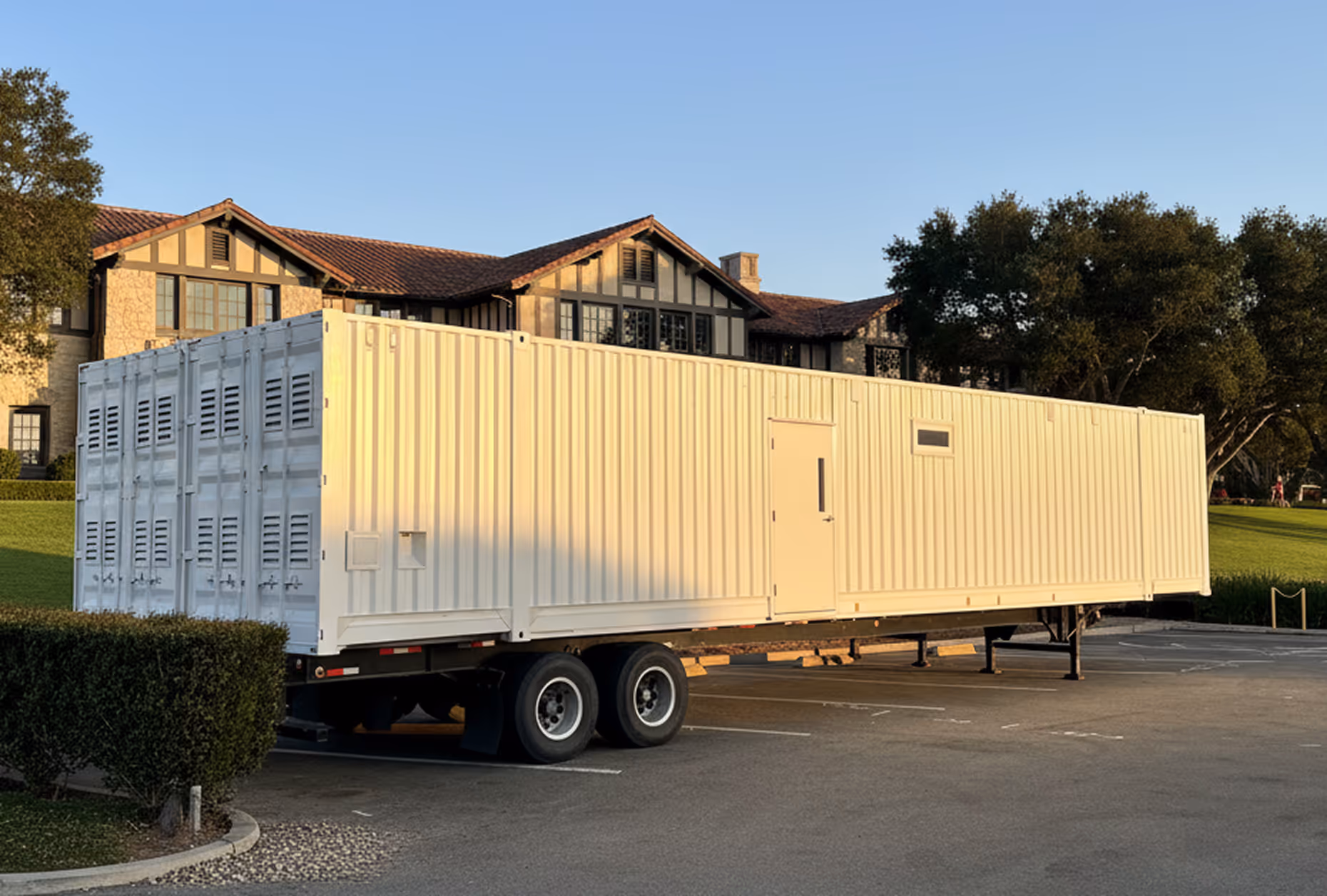 Large white mobile kitchen rental on trailer in parking lot near a building and trees.