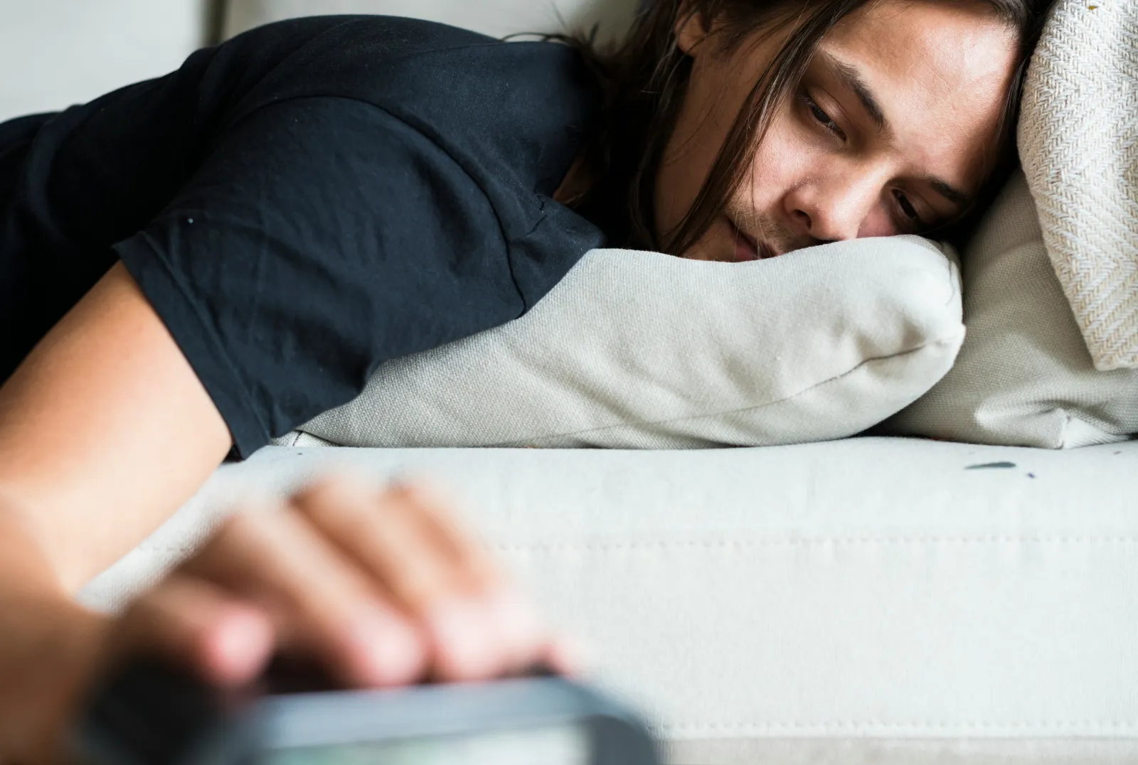 Mężczyzna leża na kanapie i sięga do budzika. Jest zmęczony i niewyspany.