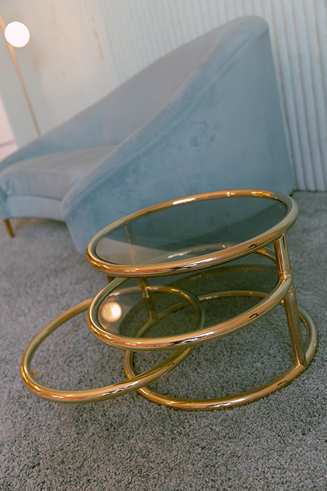 A glass table with gold details in a blue room with a blue couch in the background