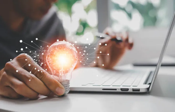 Photo of a man in front of a laptop holding a lightbulb in his right hand
