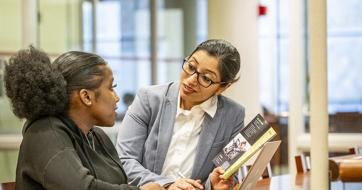 Two women in professional dress in conversation at a table inside a university building, one pointing to a laptop screen.