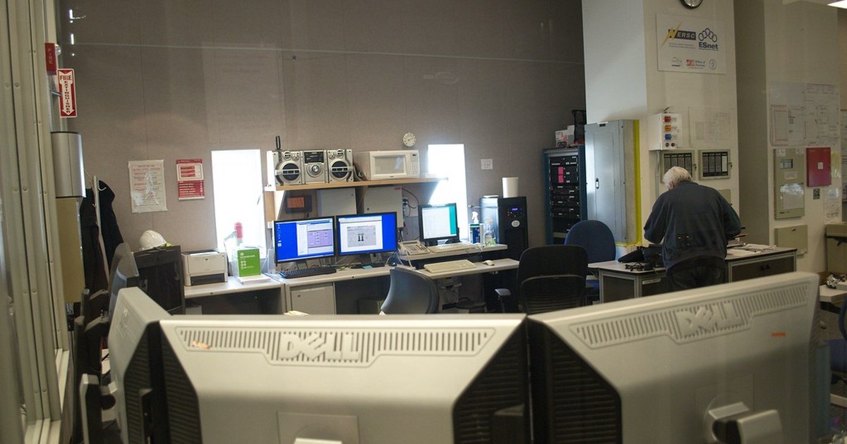 A technician monitors multiple computer screens in a data center server room while working at a workstation.