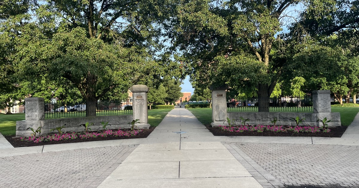 Purdue University's limestone entrance pillars line a tree-shaded campus walkway bordered by flower beds, the gateway students cross each fall semester.