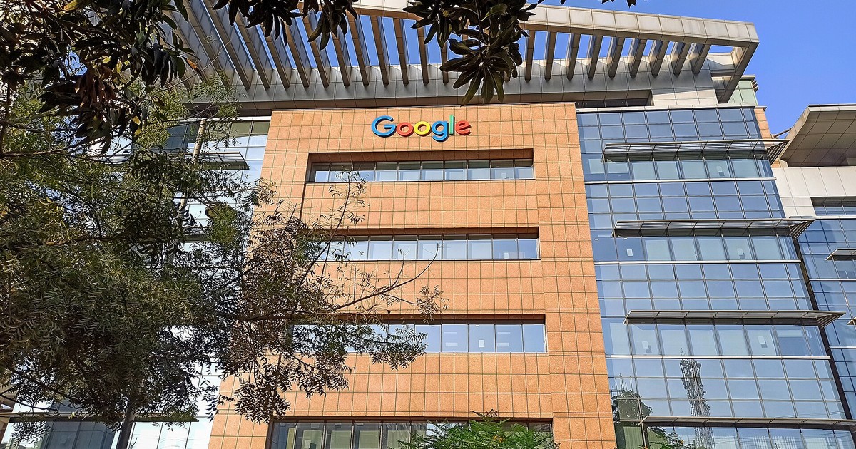 Exterior of a Google office building with the Google logo on its facade, framed by tree branches against a blue sky.
