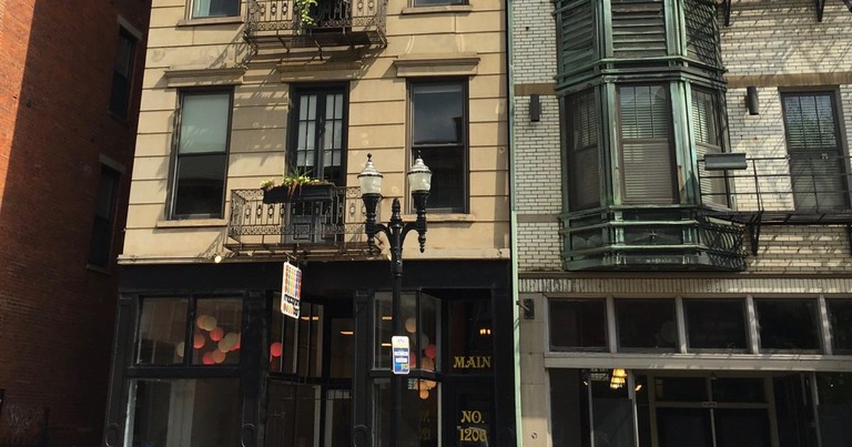 A late-afternoon street view of a small boutique storefront with black-framed windows and gold lettering, beneath an ornate apartment facade and an antique street lamp.