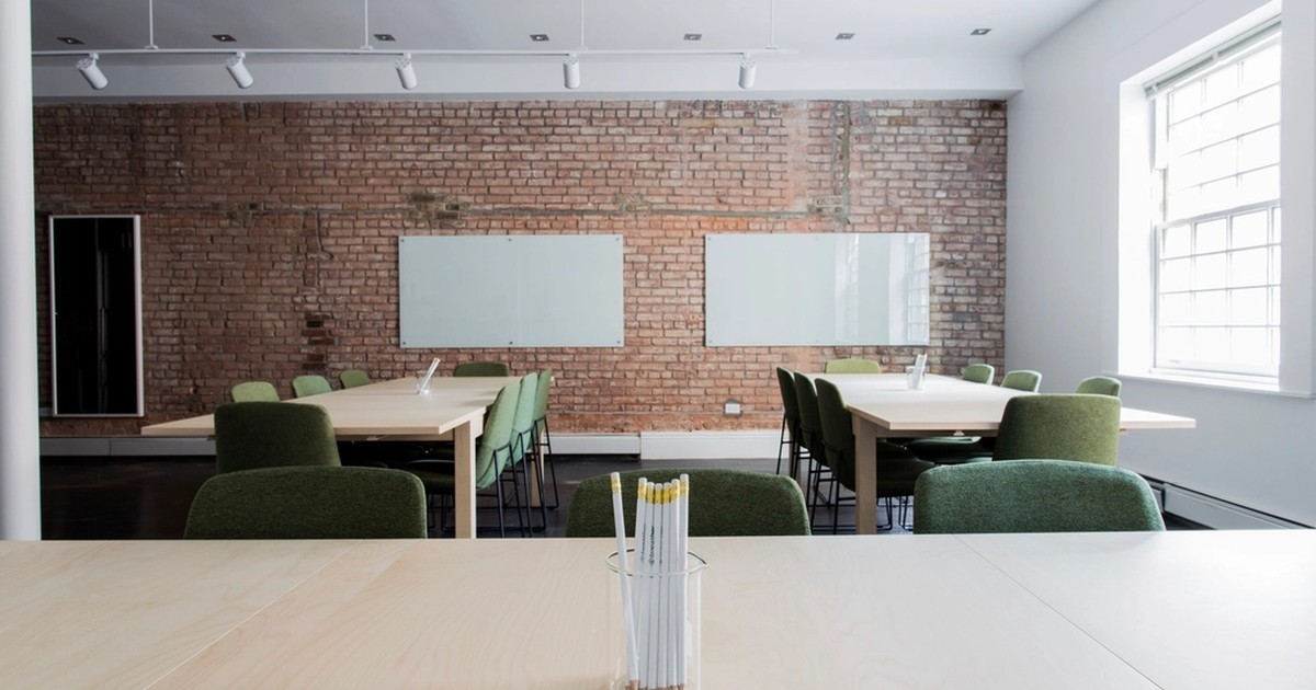 An empty, loft-style office meeting space with an exposed brick wall, two whiteboards, and green chairs pushed in around bare wooden tables, with a cup of pencils sitting on a table in the foreground.