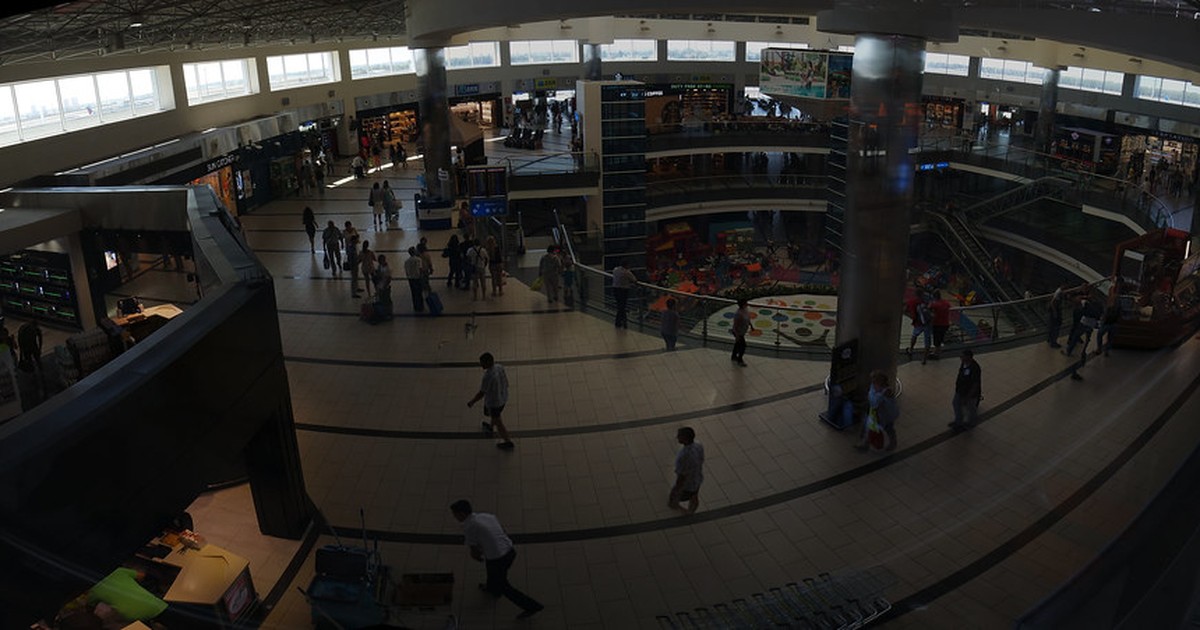 An aerial view inside a busy international airport terminal, with travelers walking past shops, information screens, and an escalator beneath a glass ceiling.