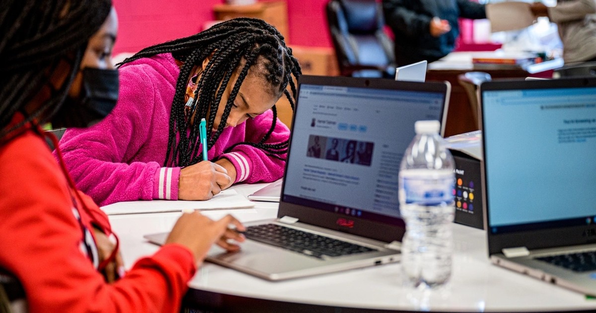 Two students at a table doing schoolwork with laptops open, one writing by hand on paper while checking the screen.