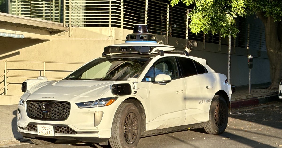 A white Waymo self-driving vehicle with a roof-mounted sensor array parked on a city street in daylight.