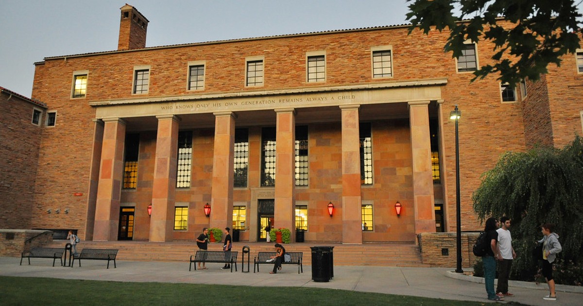 The University of Colorado Boulder's Norlin Library at dusk, its columned entrance lit and students gathered outside on the plaza.