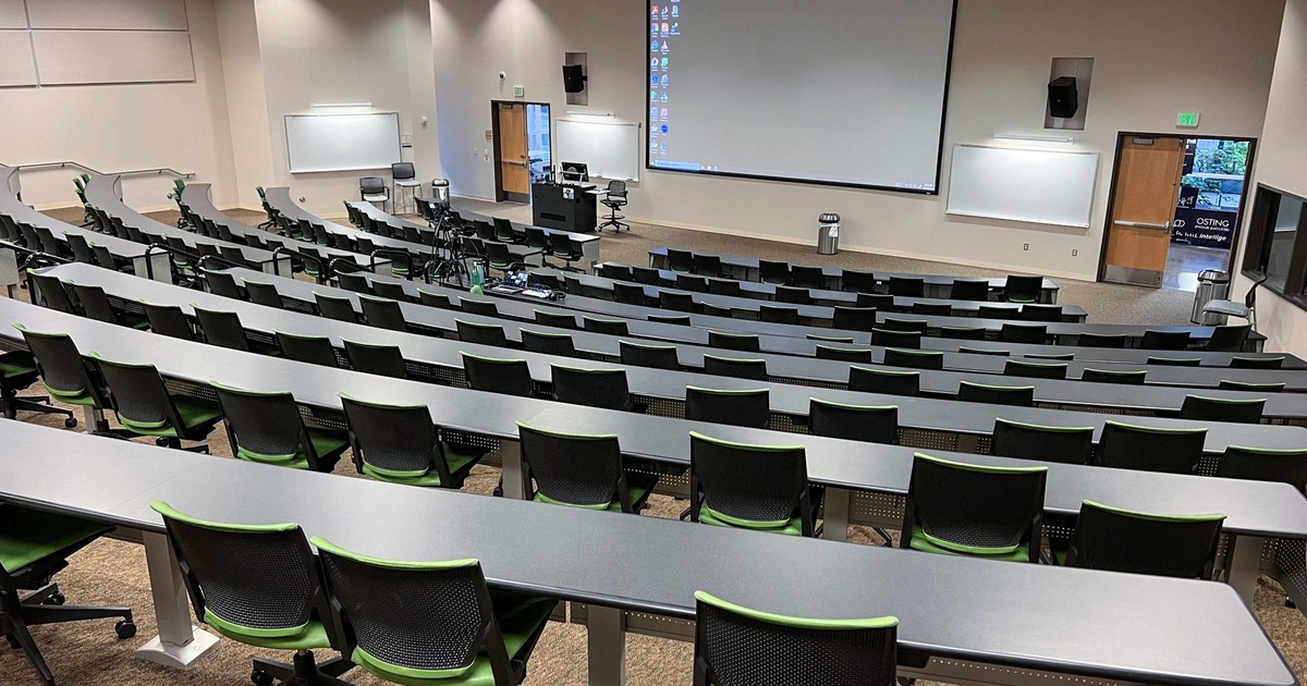 An empty college lecture hall with rows of tablet-arm chairs facing a large projector screen, the kind of classroom now covered by Florida's proposed AI policy rule.