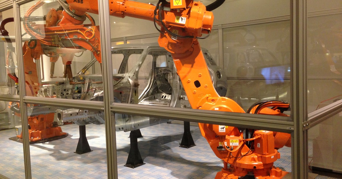 An orange ABB industrial robot arm welds a car body frame inside a glass display case at a technology museum, the kind of factory automation at the center of Bill Gates' robot tax proposal.