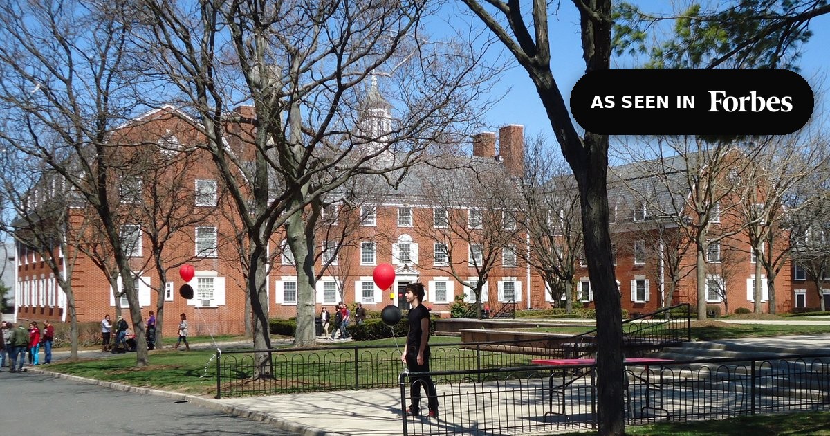 Students crossing a red-brick university campus quad in early spring, with a colonial-style academic hall behind bare trees, marked "As Seen In Forbes."