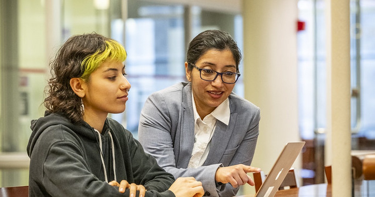 A college student sits at a library table working on a laptop with an open notebook and textbook beside them, the everyday coursework setting where AI cheating cases now begin.