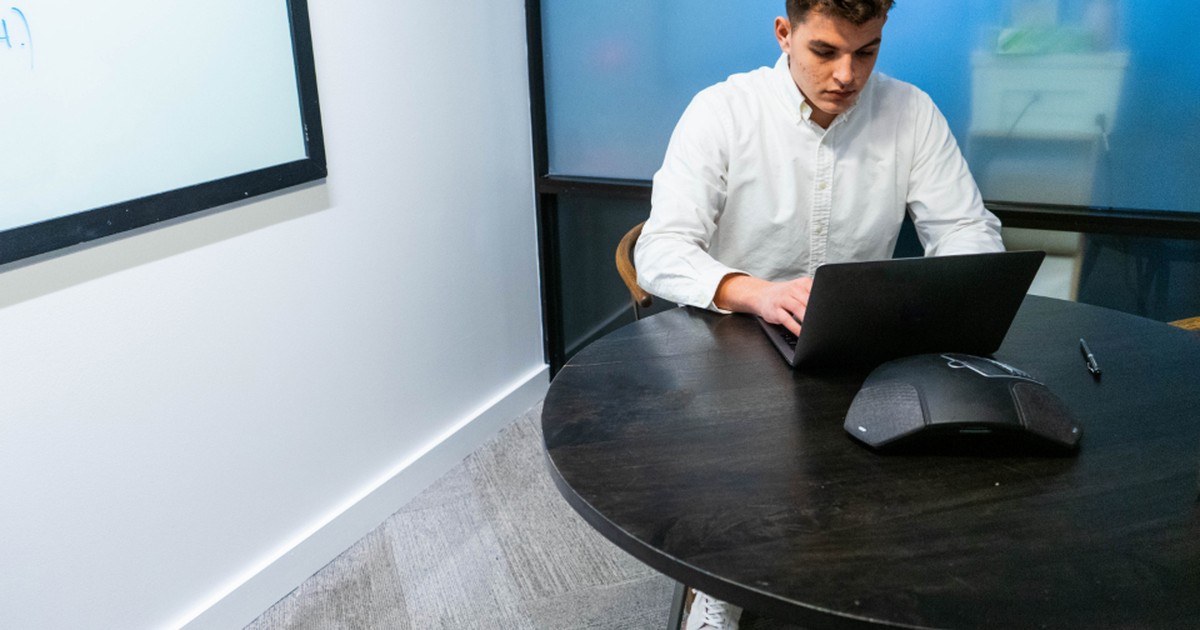 A young professional working on a laptop at a table in a modern glass-walled office meeting room, with a video-conferencing device on the table.
