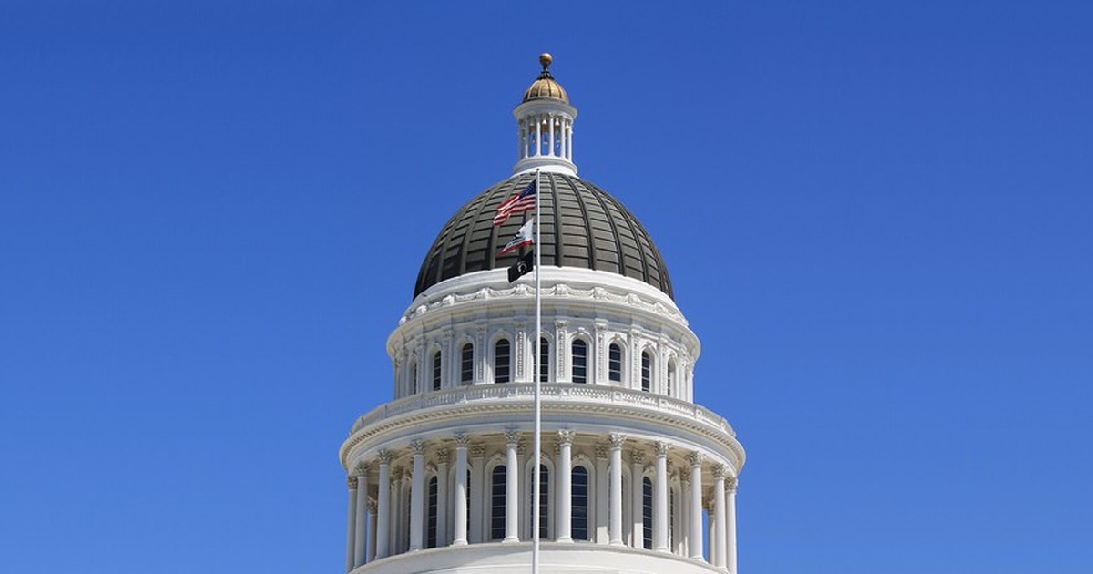 The California State Capitol dome in Sacramento against a clear blue sky, with the American and California state flags flying beside it.