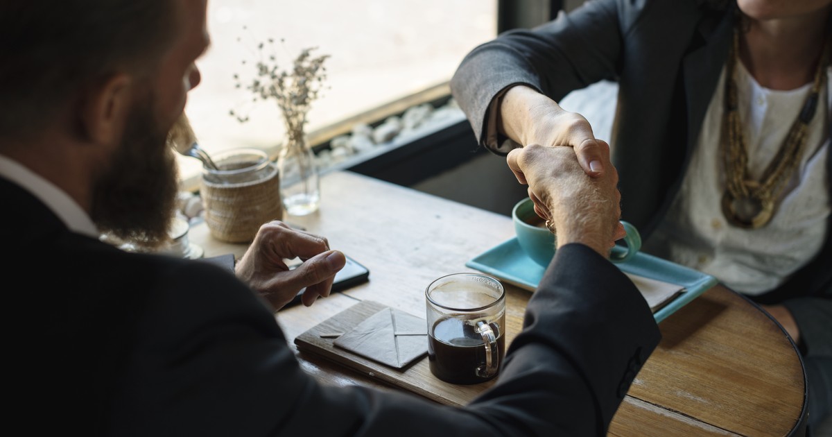 Two business people in suits shaking hands across a cafe table set with coffee, symbolizing a signed agreement between companies.