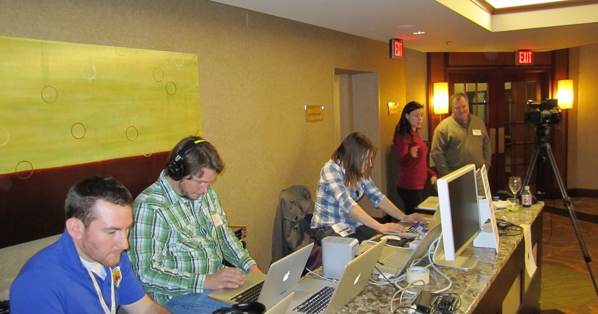 People working at laptops and monitors in a media production workspace, with a video camera on a tripod in the background.