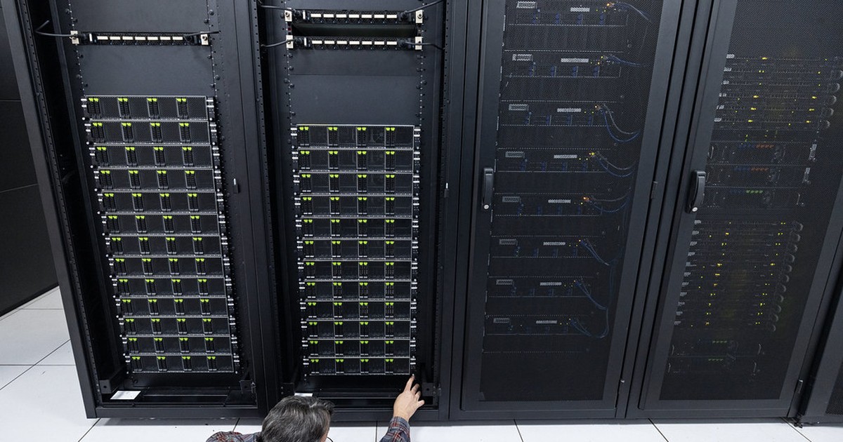 A technician kneeling beside open server racks in a data center, adjusting cabled computing hardware.