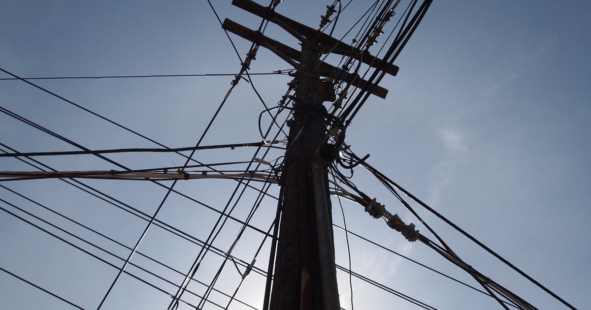 A wooden telephone and utility pole crowded with overhead wires and cable junctions, photographed from below against a clear blue sky.