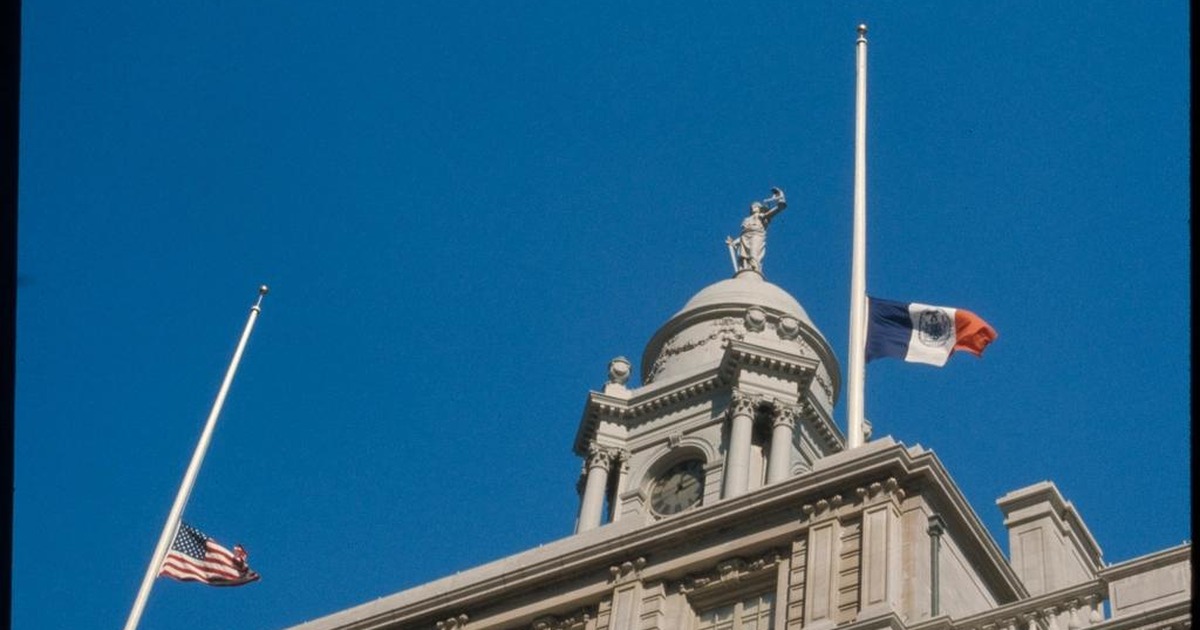 New York City Hall's cupola, topped by a statue of Justice, with the city and national flags flying below against a clear blue sky.