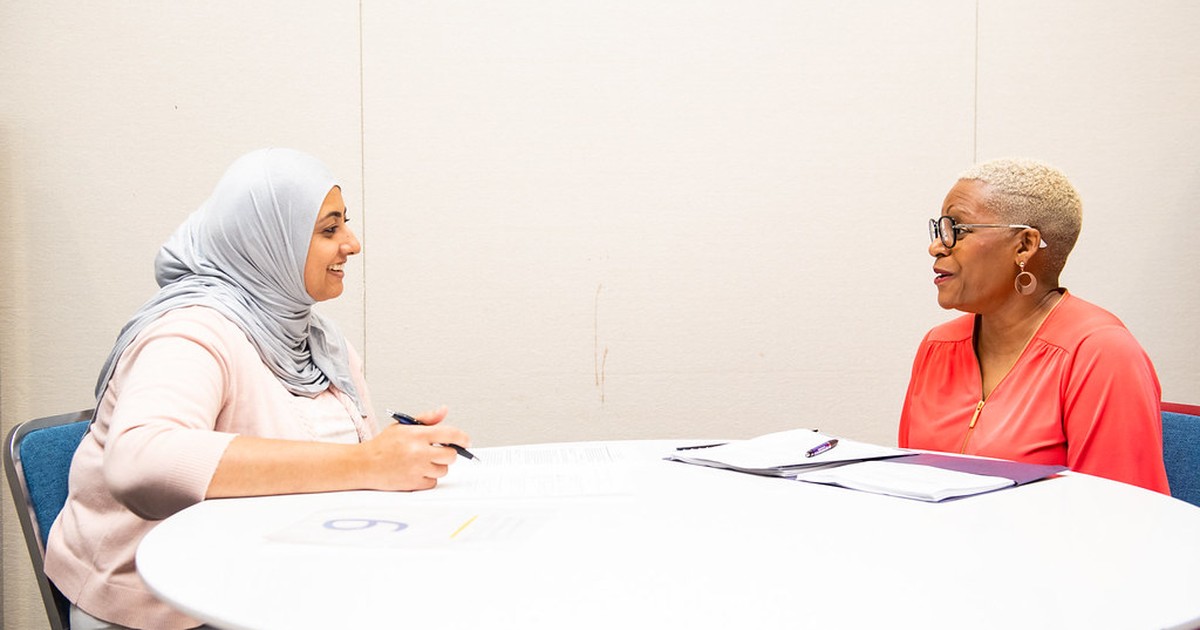 Two women sit across a table in conversation at a public job fair, one reviewing paperwork with a pen in hand.