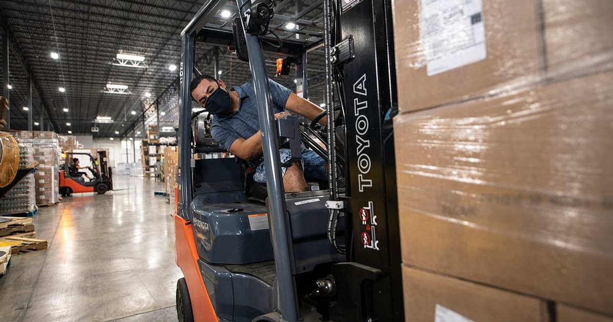 A masked warehouse worker operates a forklift near stacked boxes in a distribution warehouse.