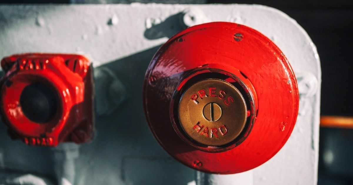 A close-up of a red industrial emergency-stop button with a brass center plate embossed PRESS HARD, mounted on a painted metal panel next to a smaller red valve fitting.