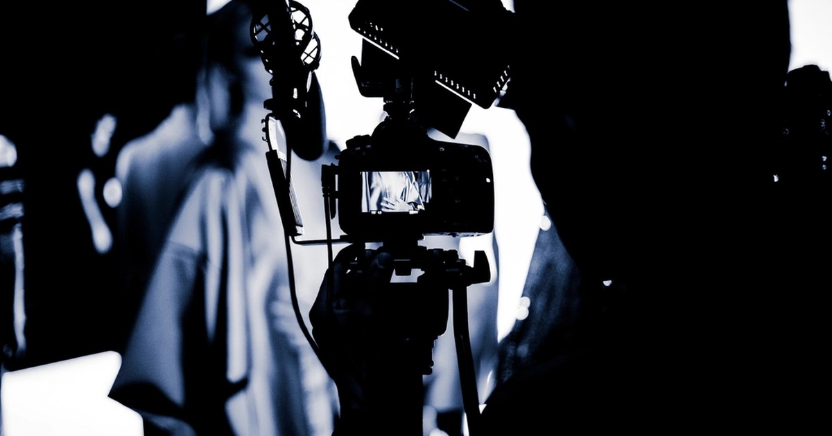 A video camera on a tripod with a boom microphone, silhouetted against bright studio light during an ad production shoot.