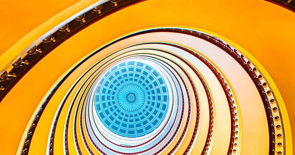 Looking straight up through a golden spiral staircase toward a round skylight, its rings narrowing toward the light.