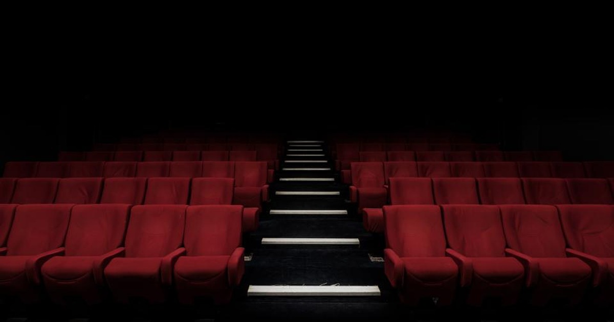 Rows of empty red auditorium seats facing a dark stage, with aisle lights leading down the center.