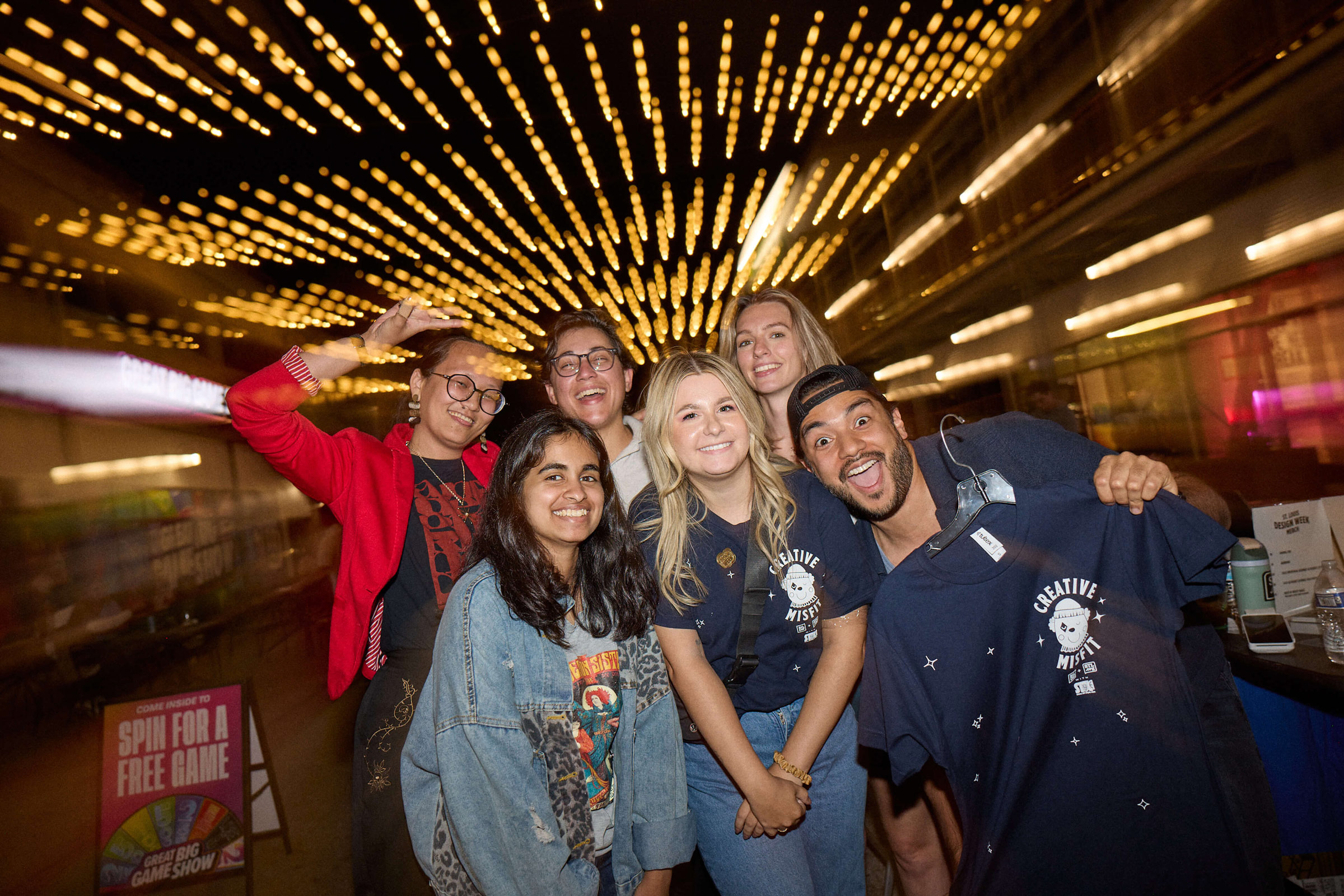Group of six young adults smiling and posing together under a ceiling decorated with warm string lights at a nighttime Design Week event.