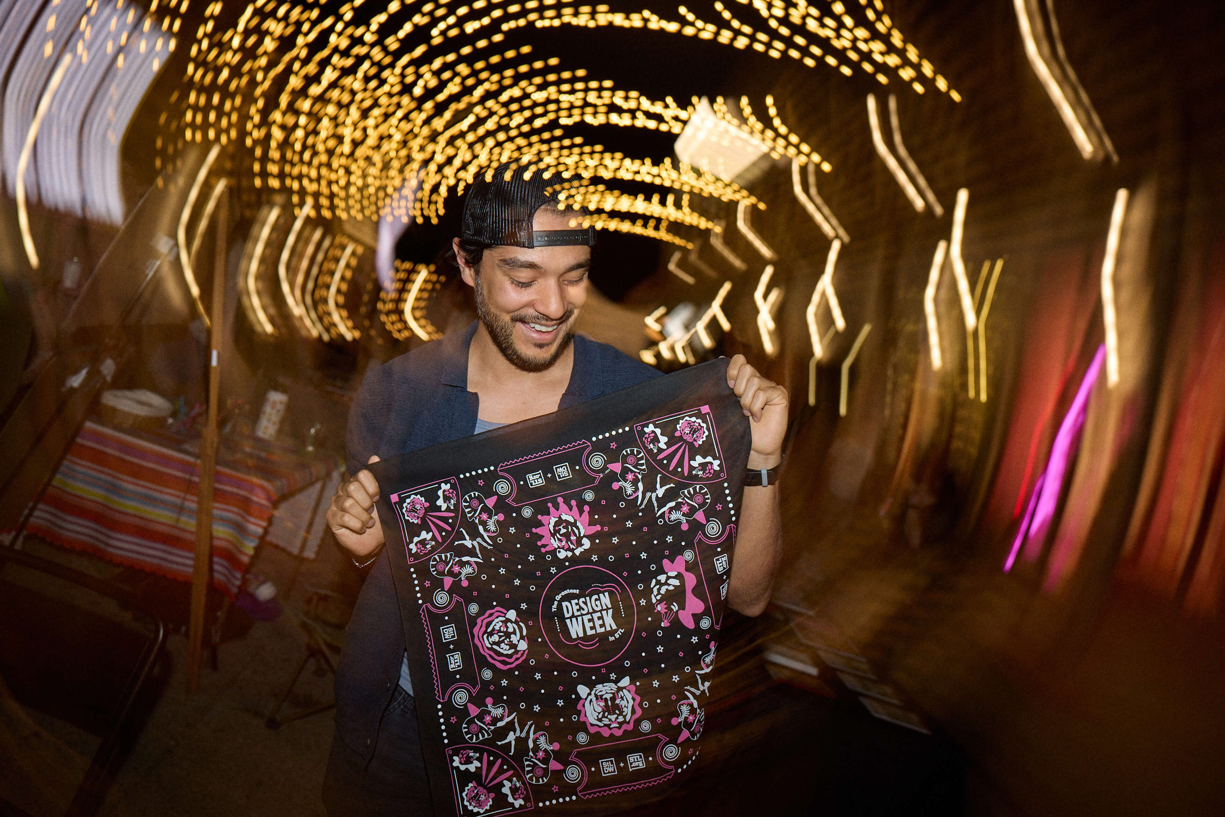 Man wearing a backwards cap smiling and holding a black cloth with colorful designs and the words 'Design Week' at night with blurred string lights in the background.