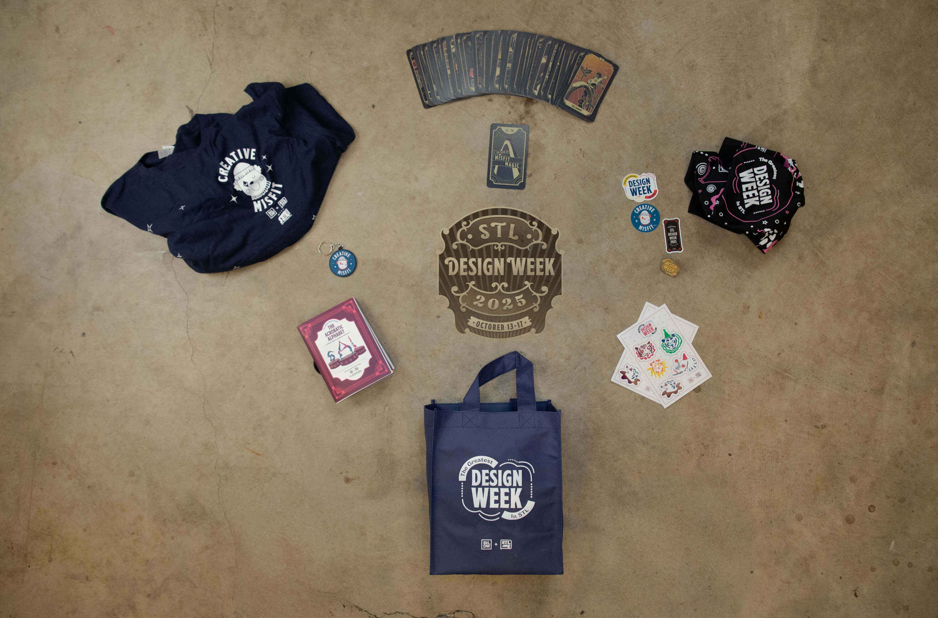 STL Design Week 2025 swag including navy tote bag, t-shirts, sticker sheets, keychain, playing cards, and a card deck on brown floor.