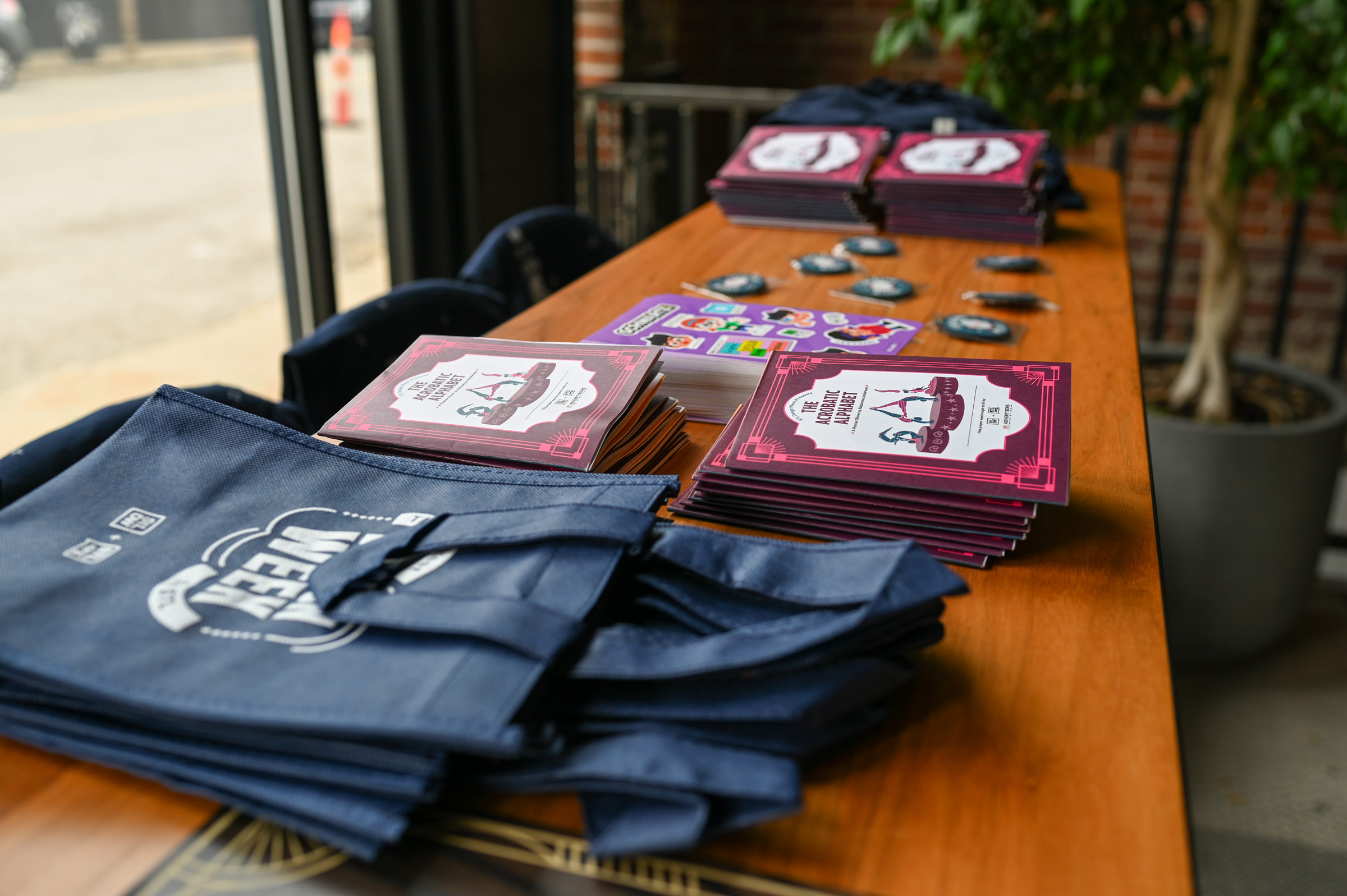 Stacks of pamphlets titled 'The Acrobat Alphabet' and blue tote bags arranged on a wooden table near a window.
