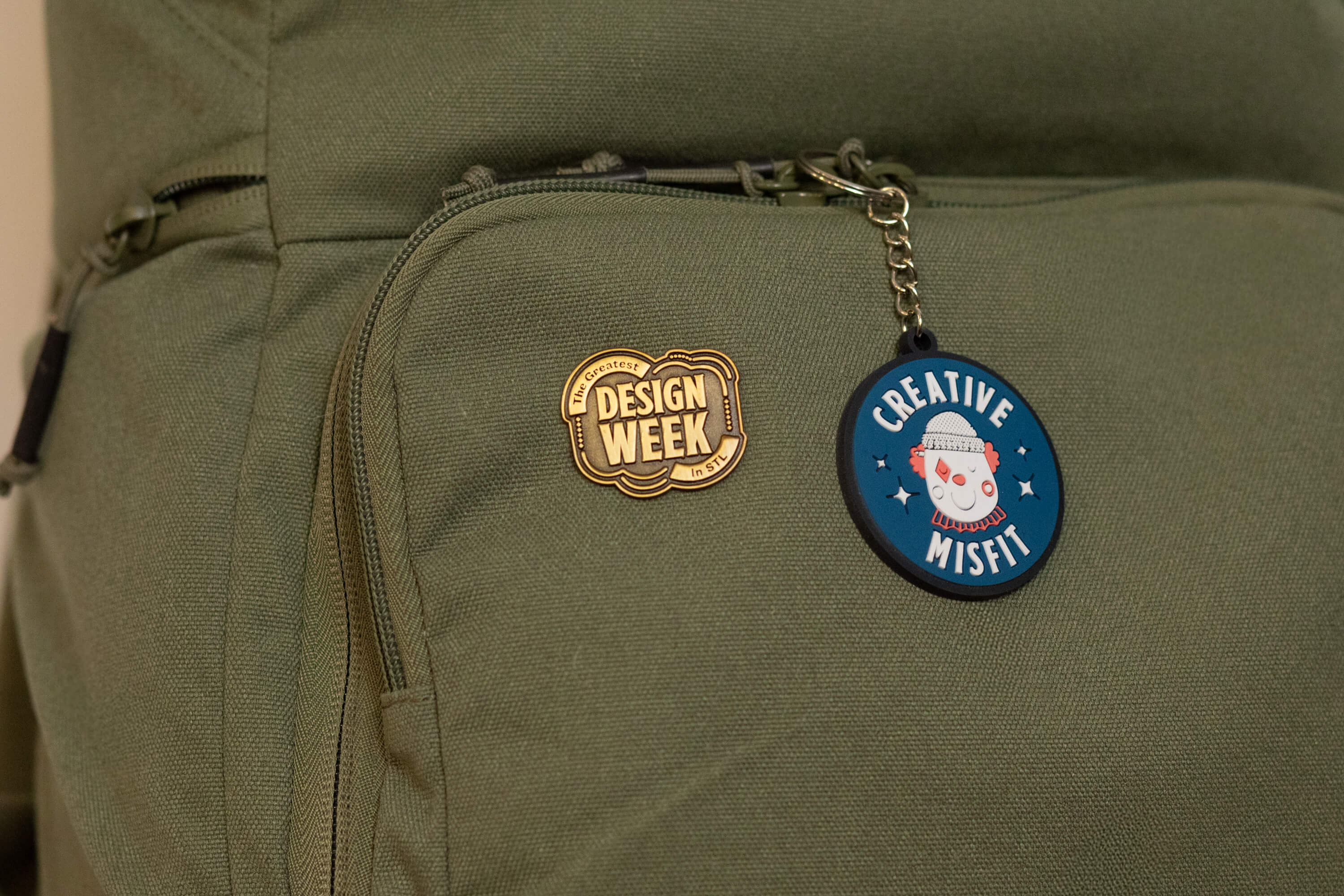 Olive green backpack featuring a gold enamel pin reading 'The Greatest Design Week in STL' and a blue round keychain with a clown face and the words 'Creative Misfit'.